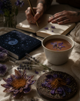 Person writing in a journal with a cup of cacao, blue lotus flowers, and crystals on a table.