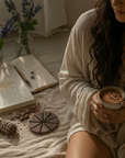 Woman in a robe holding a cup of cacao on a bed with books and flowers.