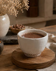 A white ceramic cup filled with cacao sits on a round wooden coaster on a rustic wooden table, with a lit candle, dried flowers, and a small bowl of cacao seeds in the background.