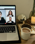 Laptop with a video call open, surrounded by two cups of cacao, a candle, and decorative items.
