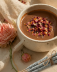 Cup of cacao with dried roses and copal sticks on a textured surface with a pink flower and candles.