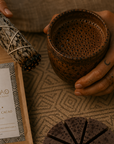 Person holding a cup of cacao with a sage bundle and a Herbal Cacao packaging on a textured surface.