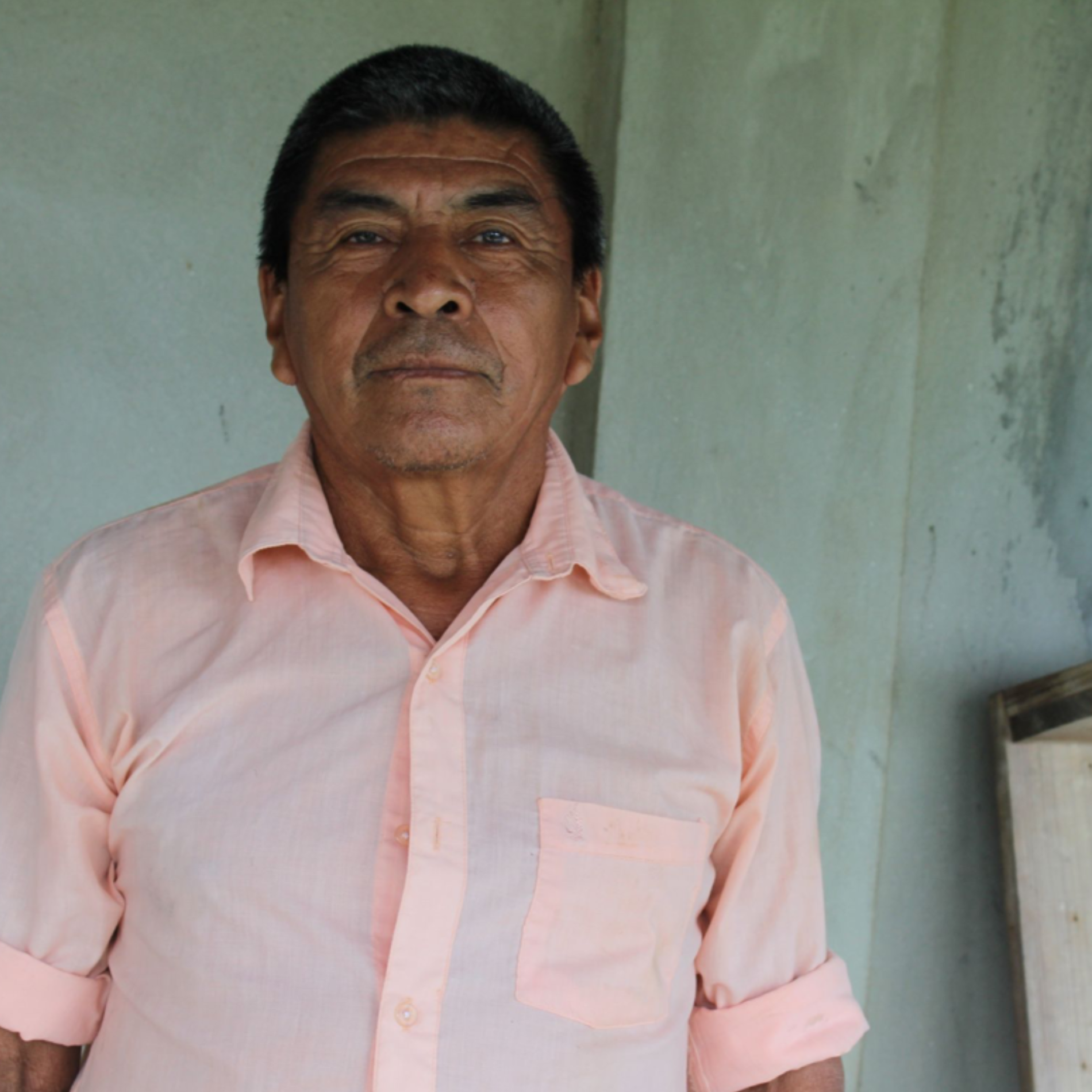 Man wearing a light pink shirt standing against a plain wall. Cacao_Farmer_Maya_Mountain
