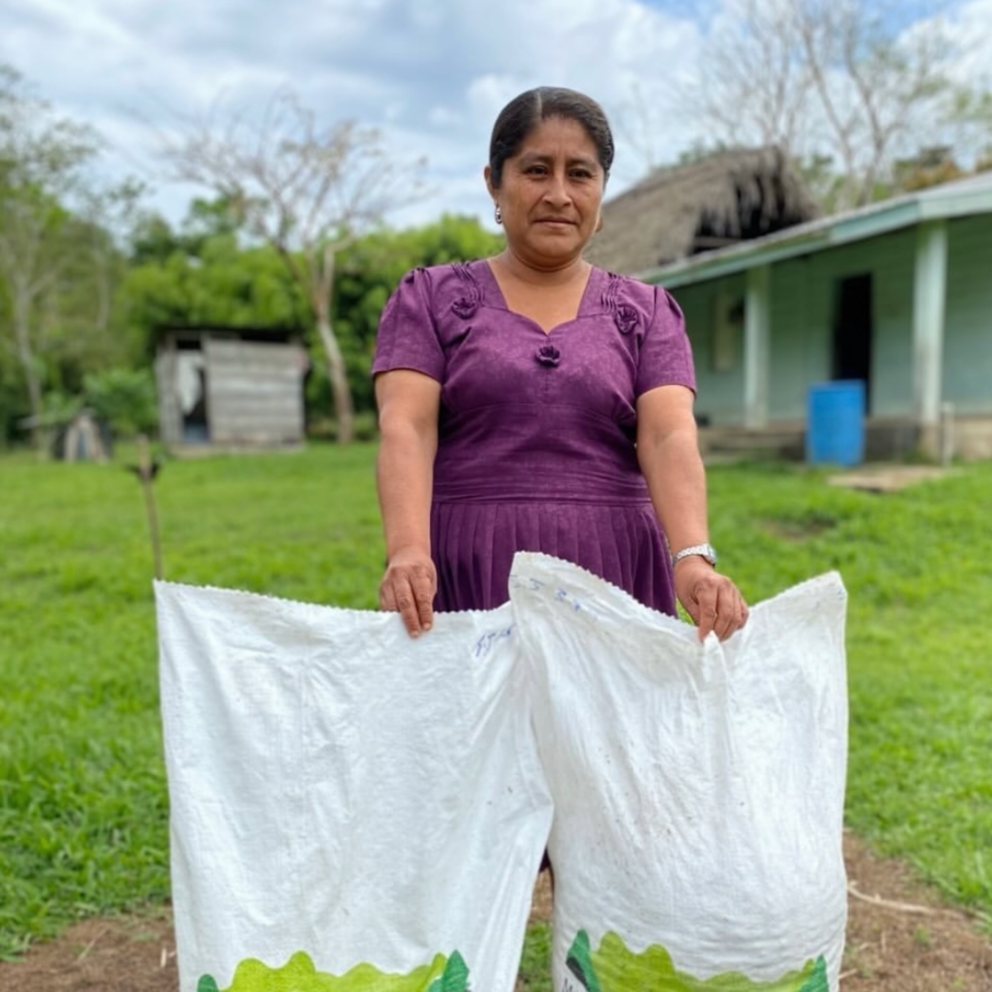 Woman holding two white cacao bags with green text in a rural setting. Cacao_Farmer_Maya_Mountain