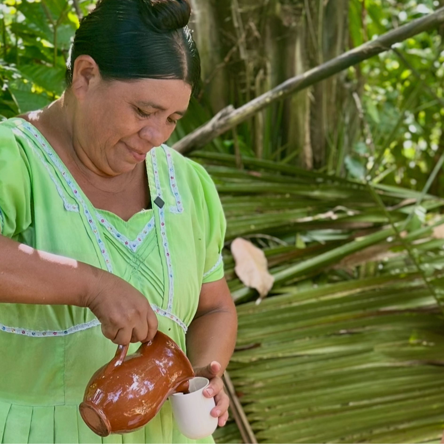 Cacao_Farmer_Maya_Mountain women in a green dress pouring from a ceramic jar into a white cup outdoors.