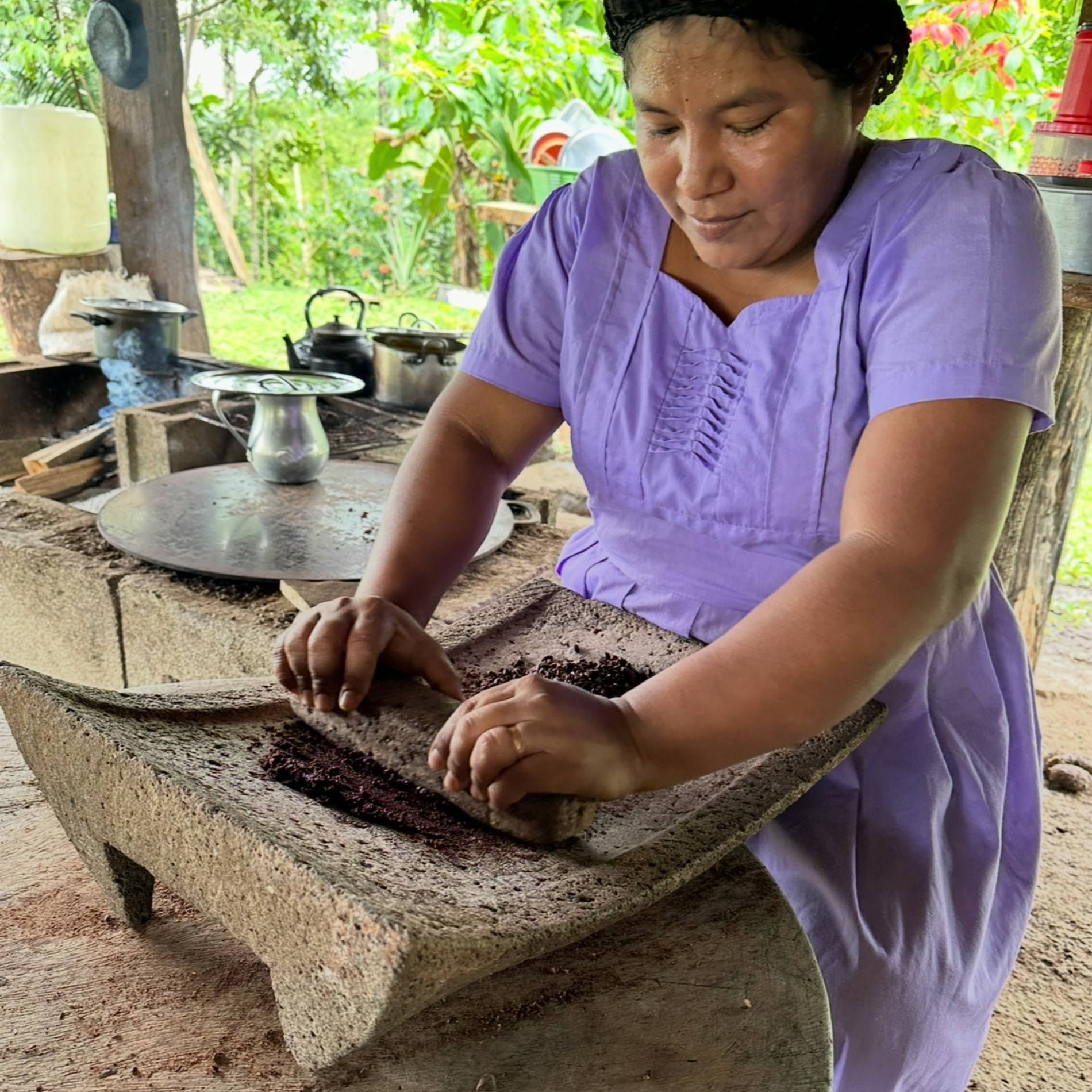 Cacao_Farmer_Maya_Mountain women in a purple shirt using a stone grinder outdoors