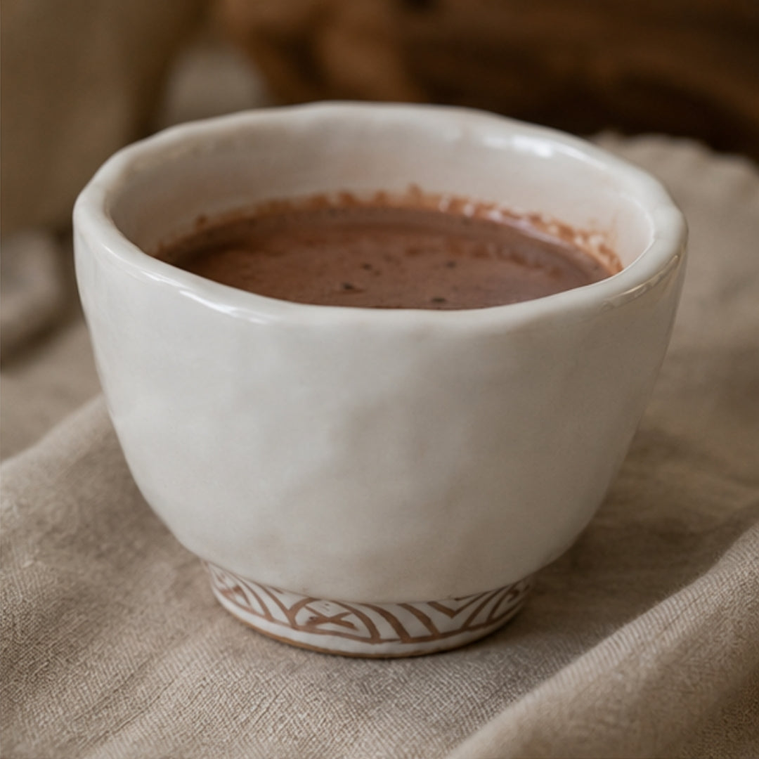 A handmade ceramic cup filled with ceremonial cacao, resting on a textured beige linen cloth with a warm, softly blurred background.