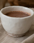 A handmade ceramic cup filled with ceremonial cacao, resting on a textured beige linen cloth with a warm, softly blurred background.