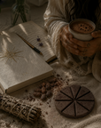 Person holding a cup of ceremonial cacao close to heart with a gratitude journal and a smudge stick on a textured surface.