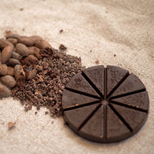 Ceremonial Cacao disc, surrounded by cacao beans and cacao nibs on a sandy surface.