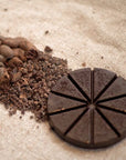 Ceremonial Cacao disc, surrounded by cacao beans and cacao nibs on a sandy surface.