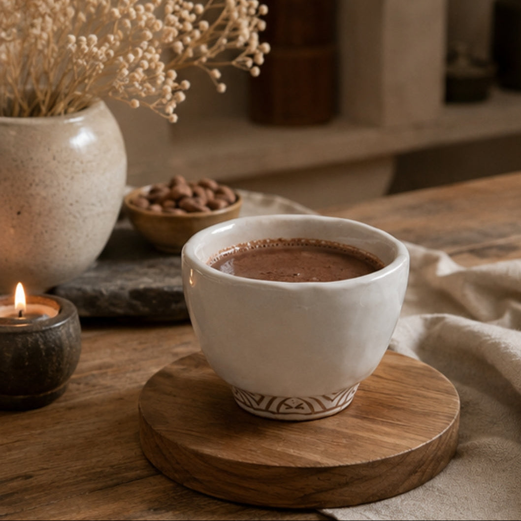 A white ceramic cup filled with cacao sits on a round wooden coaster on a rustic wooden table, with a lit candle, dried flowers, and a small bowl of cacao seeds in the background.