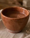 A handmade terracotta brown ceramic cacao cup with a glossy finish and a white patterned base, filled with cacao and resting on a textured beige linen cloth.