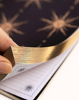 Hand holding a gold Gratitude Journal over a black surface with star-like patterns