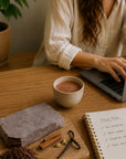Person working on a laptop with a cup of cacao, and a notebook on a wooden table.