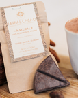 A kraft paper pouch from Herbal Cacao Natural, shown on a wooden surface with a cup of ceremonial cacao and cacao pieces nearby.