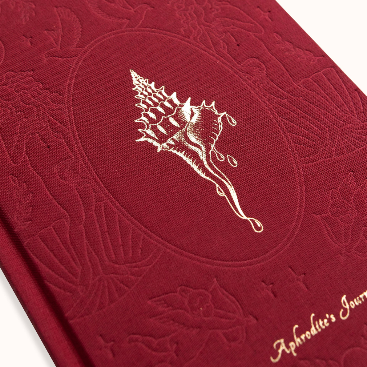Red embossed book with a shell design and 'Aphrodite's Journal' text on a white background
