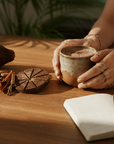 Person holding a cup of cacao on a wooden table with cocoa beans, cinnamon sticks, and a notebook.