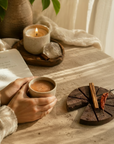 Person holding a cup of cacao, surrounded by a package of Herbal Cacao, a candle, and a book on a wooden surface.