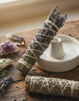 Two bundles of sage & lavender tied with twine on a wooden surface with a small white dish, crystals, and a brown ceramic bowl.