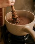 whisking a pan of warm cacao with a Molinillo a traditional whisk for cacao, next to a package of Herbal Cacao.